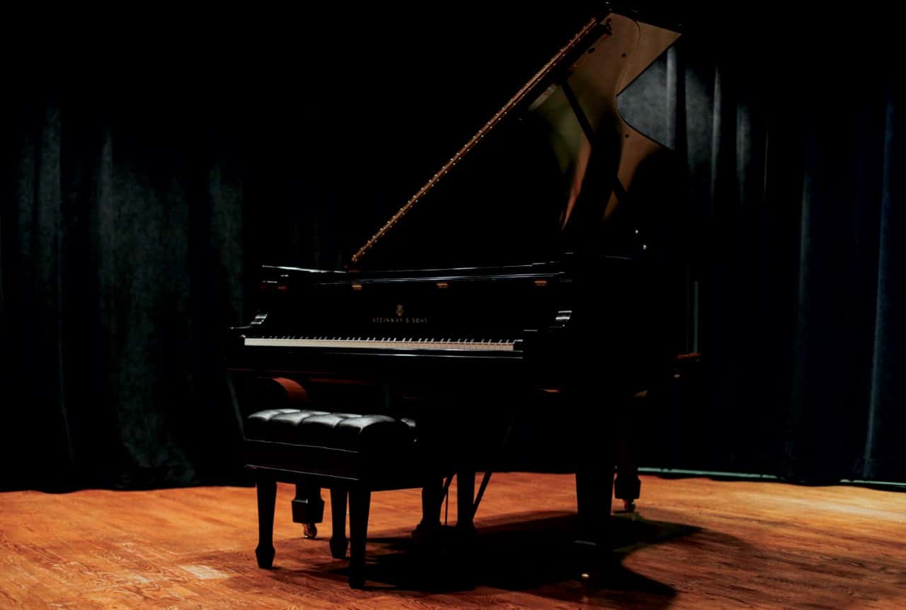 Grand piano on dimly lit stage with wooden floor and matching bench.