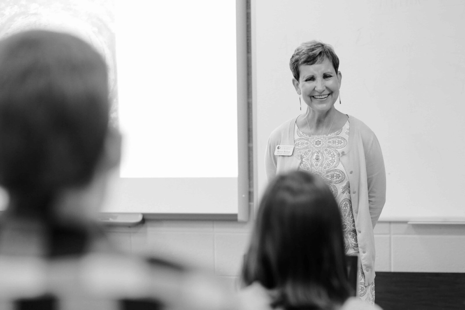 Smiling woman in patterned dress and cardigan presenting in front of a room with a screen behind.